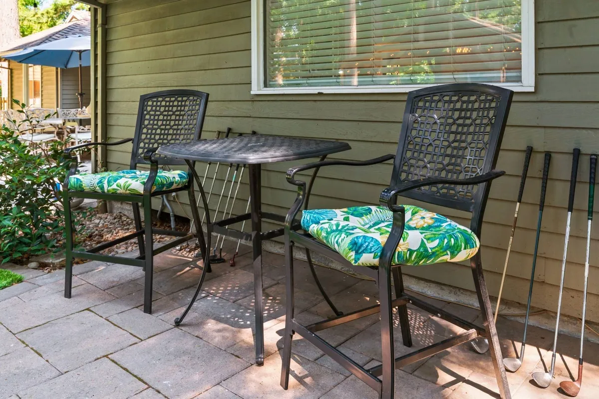 An outdoor bar-height table with two tropical-cushioned chairs sits on a patio, with golf clubs nearby. The setting evokes casual al fresco dining, shared drinks, and easy conversation against the backdrop of sunset or morning sunshine.