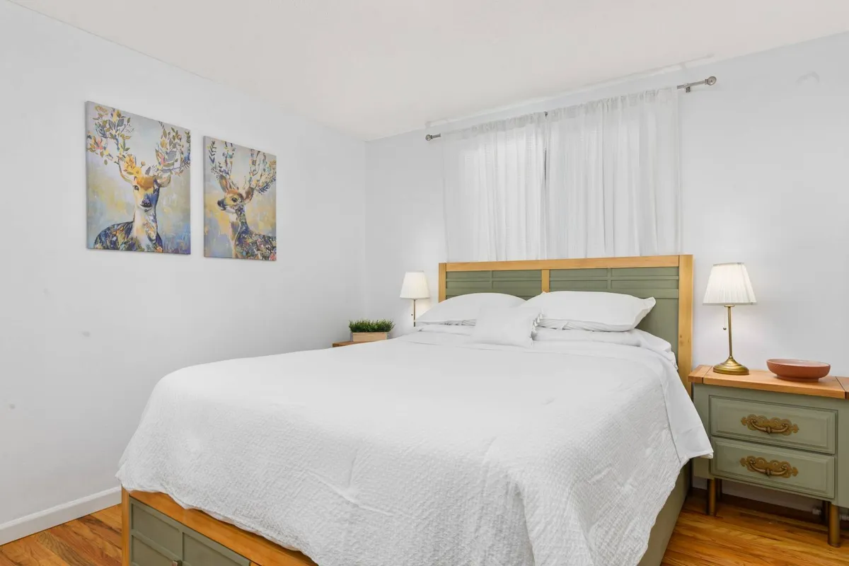 The serene bedroom features a plush bed, calming deer artwork above the headboard, and soft bedside lighting. The combination of gentle colors and inviting textures creates an atmosphere perfect for winding down and restful sleep.
