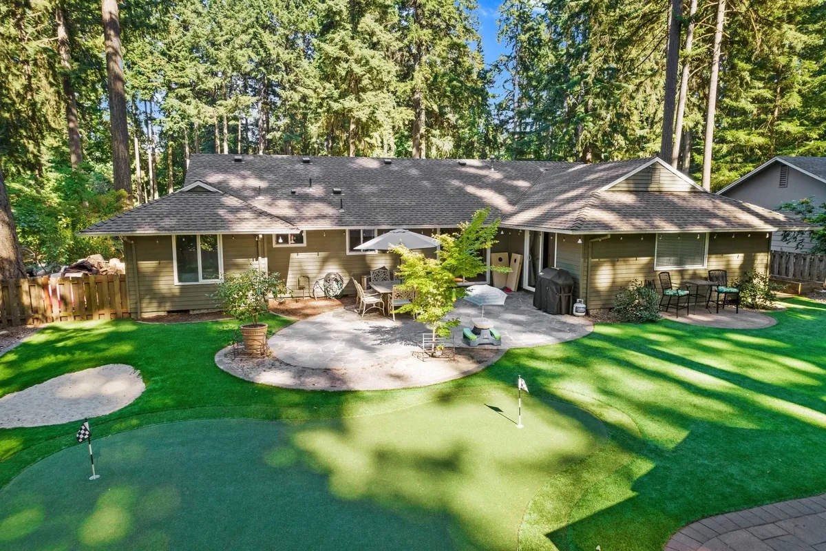 This exterior image showcases a vibrant backyard with lush green artificial turf designed as a golf putting green, a comfortable patio seating area shaded by an umbrella, and mature trees providing a tranquil setting. The space is clearly set up for golf practice as well as relaxation and family gatherings.