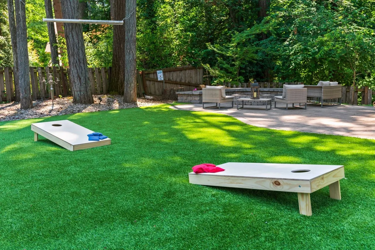 A playful outdoor scene featuring cornhole boards set up on the grass, a fire pit ready for evening gatherings, and a comfortable seating area. The space is lively yet intimate, perfect for games, socializing, and cozy nights by the fire.
