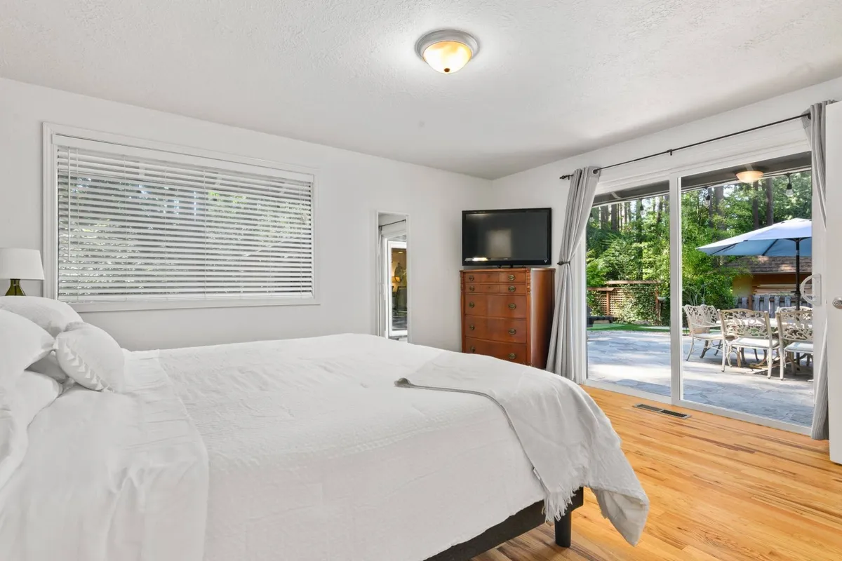 A spacious bedroom with a large bed, stylish dresser, TV, and sliding glass door that opens directly to the lush backyard. Sunlight pours through the glass, blending indoor comfort with outdoor serenity.