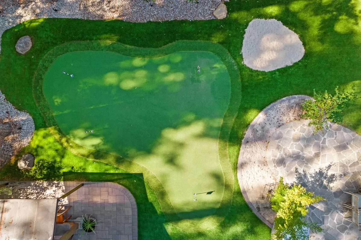 A wide view of a professionally manicured backyard featuring an artificial putting green and sand pit, surrounded by lush greenery and mature trees. There’s a spacious patio area adjacent to the green, perfect for social gatherings or relaxing after practice. The scene exudes a sense of leisure and private resort living, with visible golf equipment highlighting its specific use for golf practice.
