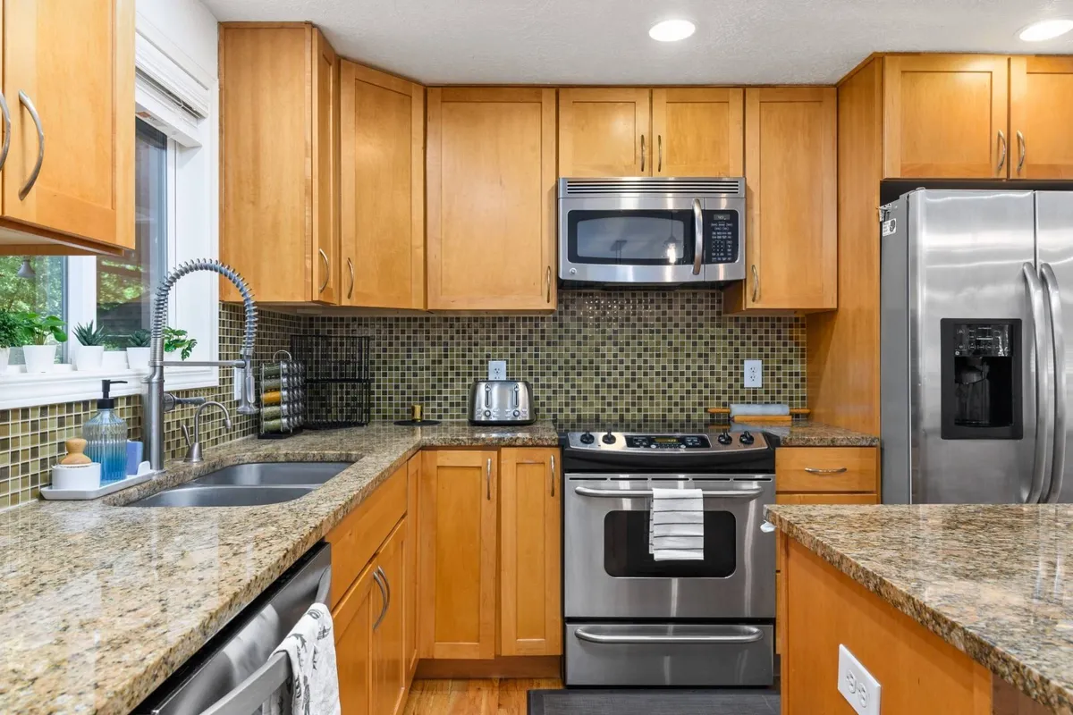 A close-up of the kitchen, highlighting the granite countertops, modern stainless steel appliances, mosaic backsplash, and wood cabinetry. The layout encourages cooking, meal prep, and interaction, with ample space for multiple people to help or chat together.