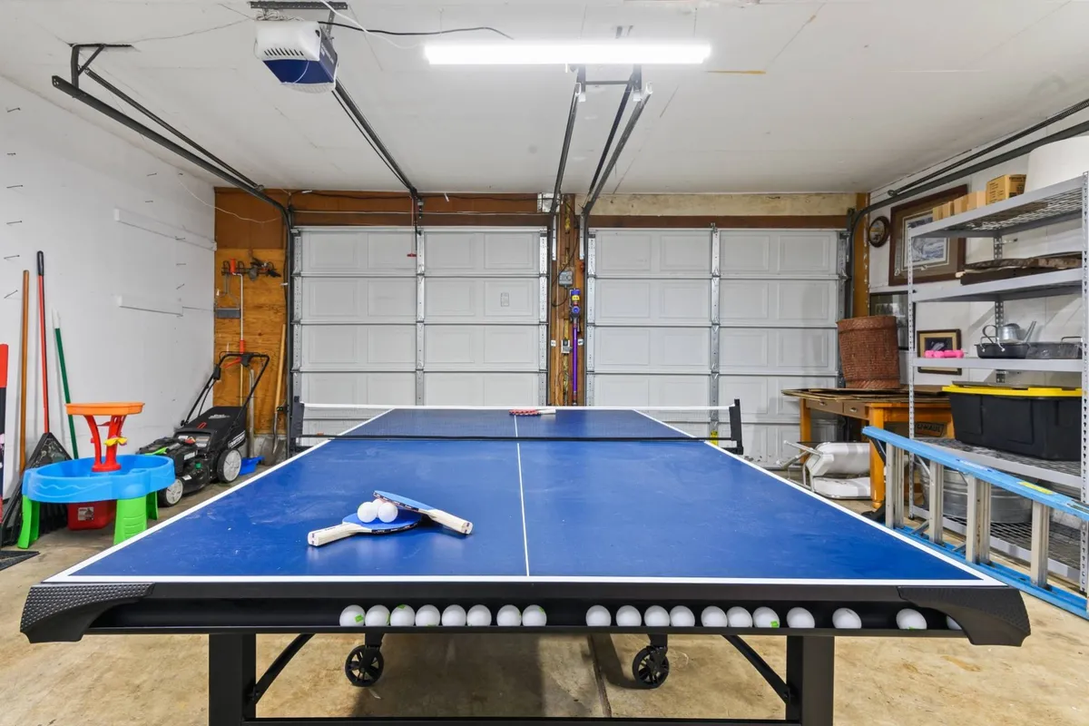 A bright and lively garage space, featuring a full-size ping pong table set up and ready for action. Table tennis paddles rest on the table, and the open garage doors in the background hint at easy access to the outdoors. The area is organized, with space for friendly games and social gatherings.