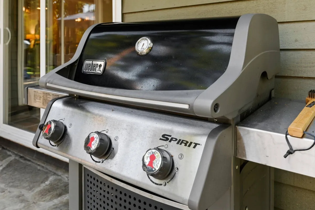 An exterior shot focusing on a modern grill situated on a spacious patio, surrounded by ample seating and reflections in nearby windows. The setup hints at an inviting environment for outdoor cooking and gatherings.