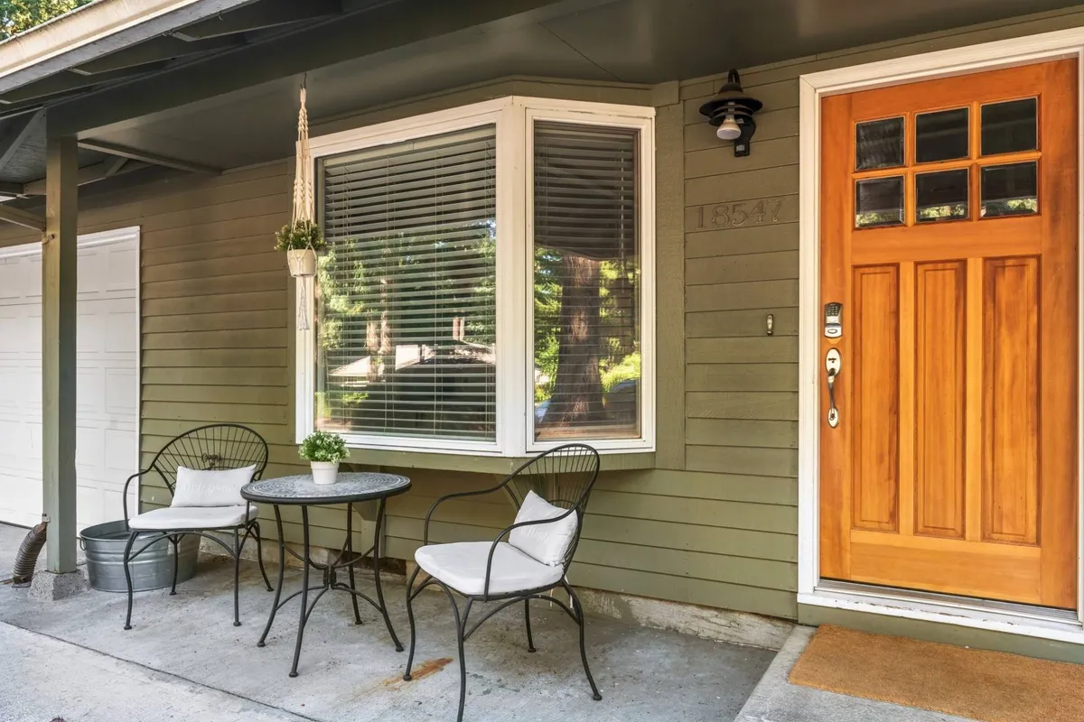 The image shows the home's welcoming front door set in a tranquil neighborhood, with a small seating area beside it and a window adorned with plants. The outdoor setup suggests a peaceful spot to enjoy morning coffee or quiet evening conversations.