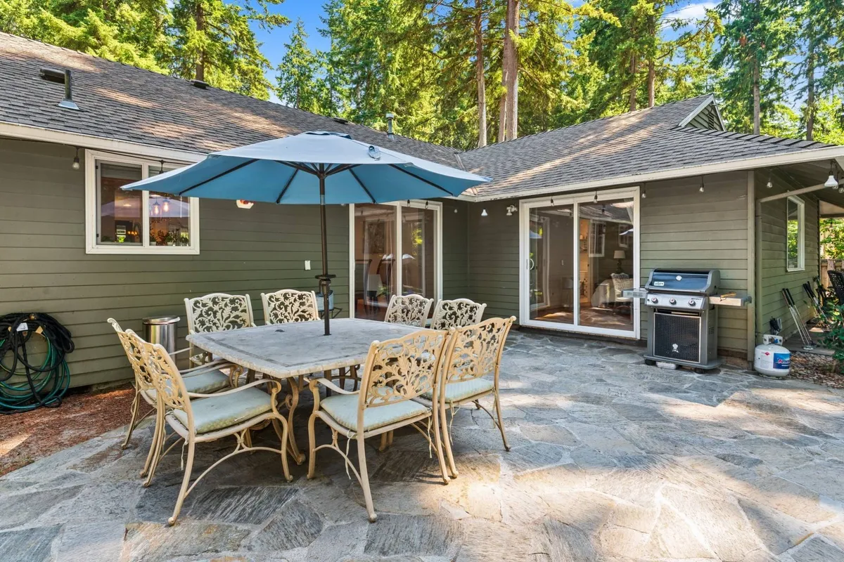 A picturesque outdoor dining area with a large table under an umbrella, a nearby grill, and lush trees in the backdrop. Sliding glass doors suggest seamless flow between indoor and outdoor living, ideal for festive group meals or tranquil evenings.