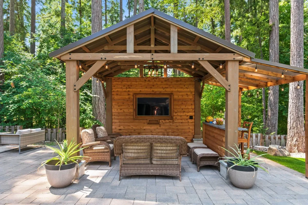 A wooden gazebo sits at the heart of the backyard, furnished with cozy wicker seating and featuring an outdoor television, surrounded by greenery. The setup invites group gatherings for entertainment, relaxation, or watching shows outdoors, sheltered from the elements.