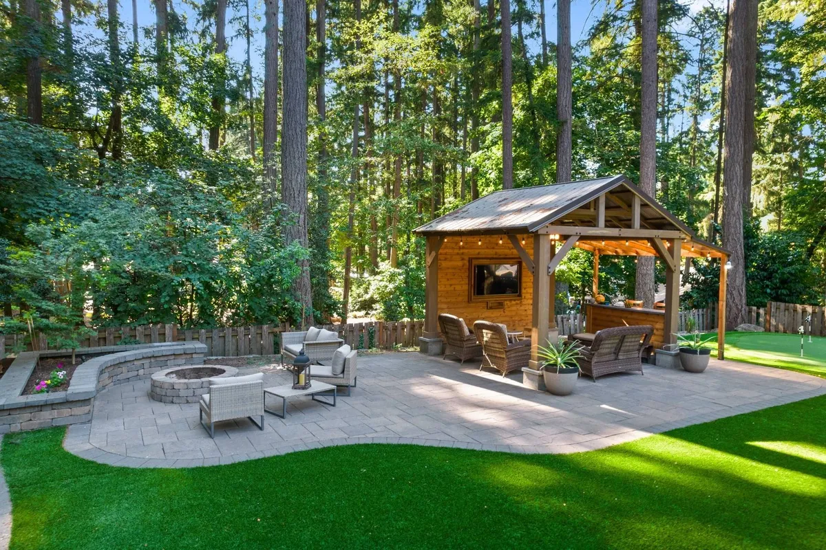 An inviting exterior space featuring a gazebo with patio seating, a stone fire pit at the center, and lush greenery surrounding the area. The ambiance is tranquil yet social, ideal for evening gatherings or winding down after a full day.