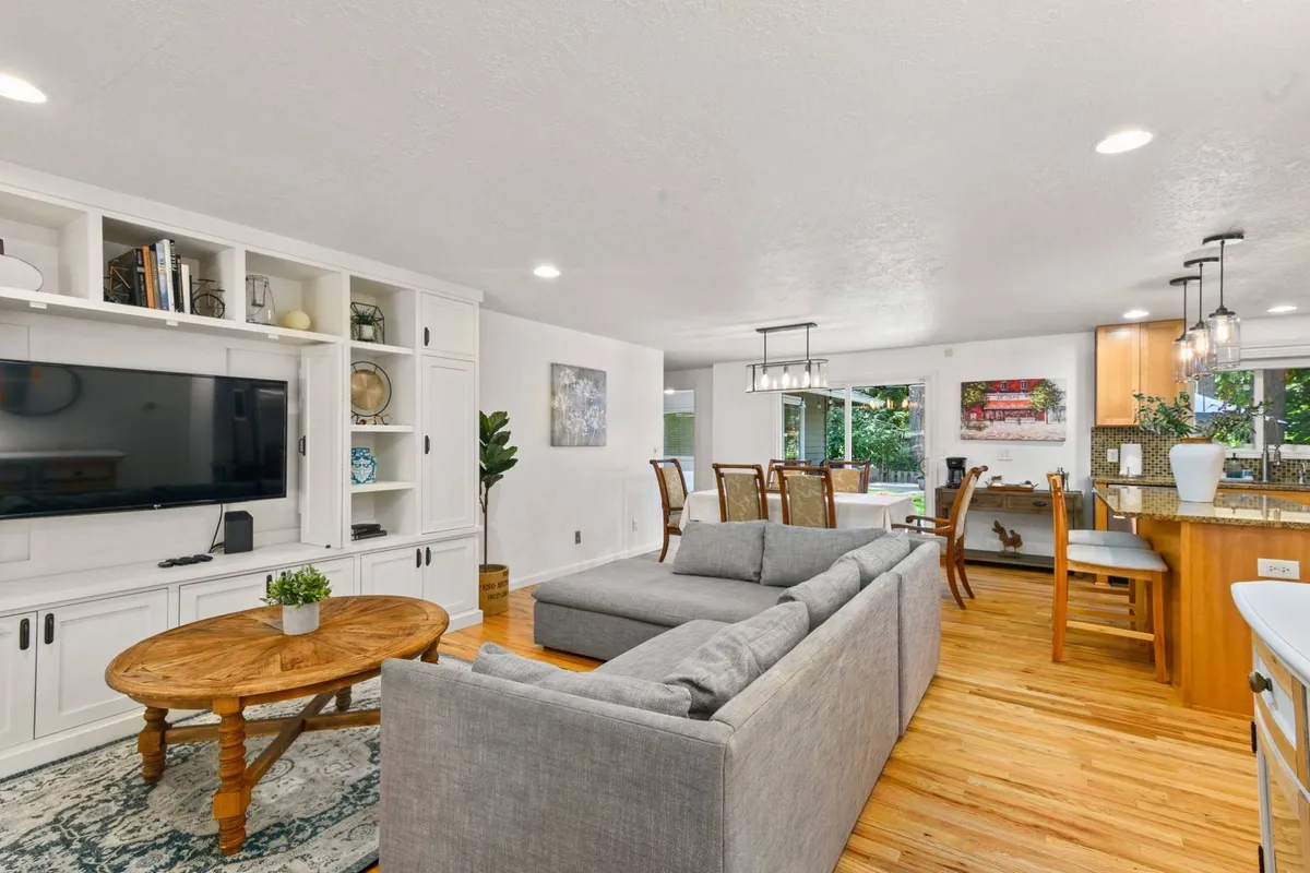 A wide view of the open-concept living space, featuring a plush sofa positioned for conversation or watching TV, a dining table ready for family meals, and a kitchen counter visible in the background, all bathed in natural light. The seamless layout highlights how living, dining, and kitchen areas are interconnected, making it easy for guests and hosts to mingle.